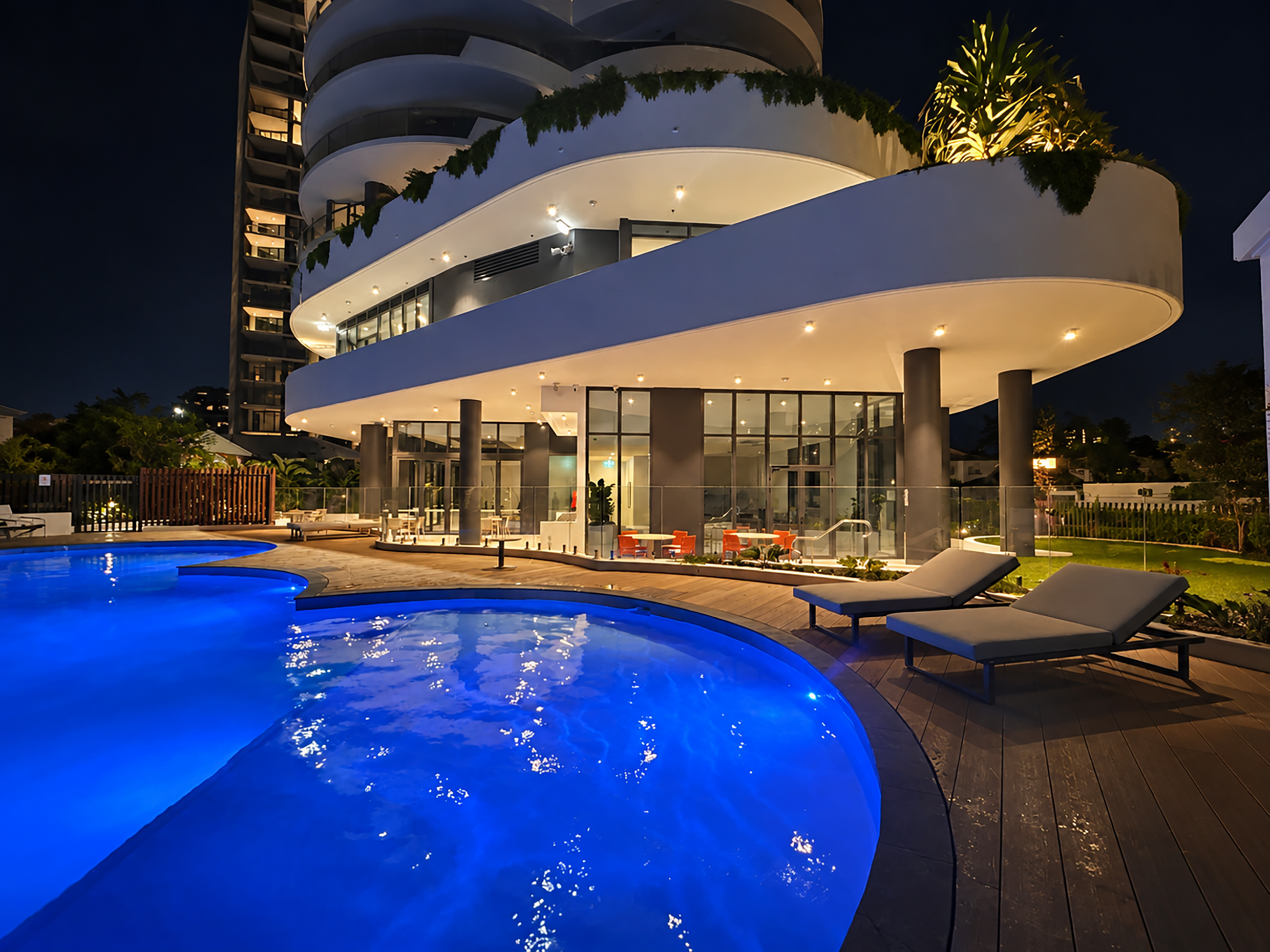 Night view of the illuminated outdoor pool and terrace at Chevron One, with sun loungers, landscaped gardens and curved apartment balconies.